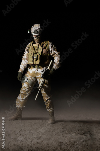 Fully equipped desert soldier standing with rifle on dark background.