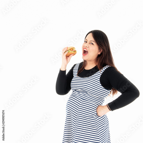 A young fat woman is eating a hamburger in her hand. She is smiling and happy to eat the food she likes. She is hungry. Healthy concept. Isolated on a white background.