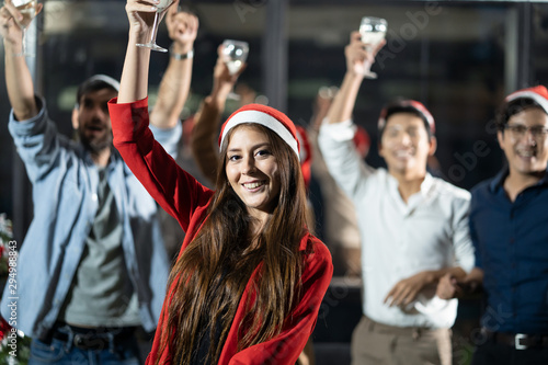 Group of various nationalities friends having Christmas party in working office at night. Caucasian pretty girl holding Champagne glass dancing in front while her colleagues having fun, happy behind. © Kawee