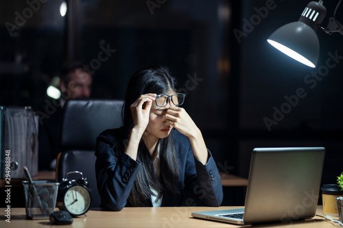Asian business woman working late sitting on desk in office at night. The girl feeling stress and tired after overload job has been assigned. Lady hold glasses up and hand on nose. Boss sits behind.