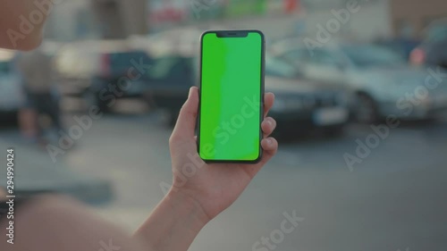 Young woman browsing content on vertical smartphone with template blank greenscreen on parking lot in shopping centre outdoors. Close-up.