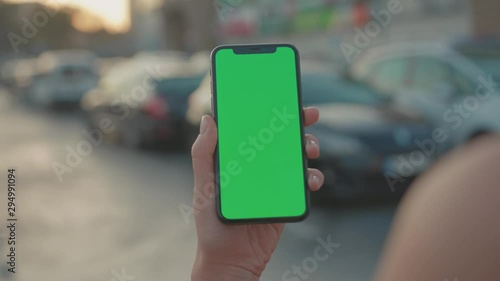 Close-up mock-up modern vertical smartphone with greenscreen in human hand. Young person holding mock-up mobile device on parking lot outdoors.