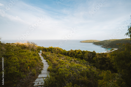 Lone woman walking down a single path through shrubs and grass towards a beautiful view of a bay at Shelley Beach, Albany, Western Australia.