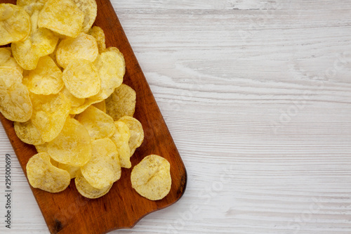 Tasty potato chips with salt on a rustic wooden board on a white wooden surface, top view. Flat lay, overhead, from above. Space for text.