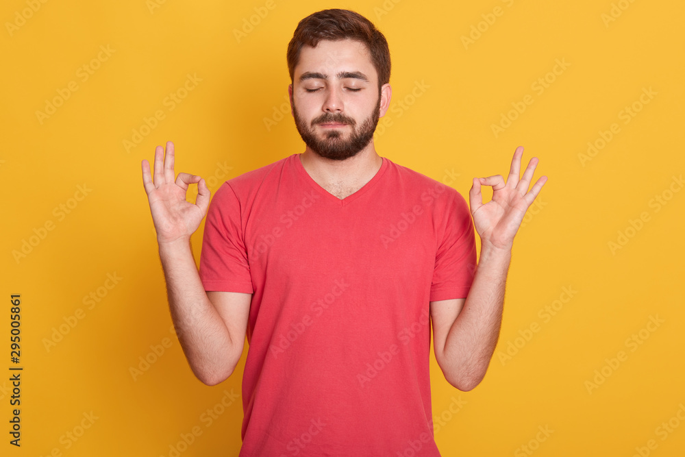 Close up portrait of attractive man dresses red casual t shirt gesturing ok sign with closed eyes and both hands , model posing isolated over yellow background in studio. Meditation concept. Close up portrait of attractive man dresses red casual t shirt gesturing ok sign with closed eyes and both hands , model posing isolated over yellow background in studio. Meditation concept.
