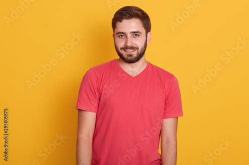 Portrait of young happy attractive bearded guy, handsome male wearing red casual t shirt, smiling and looking directly at camera isolated over yellow studio background, having calm facial expression.
