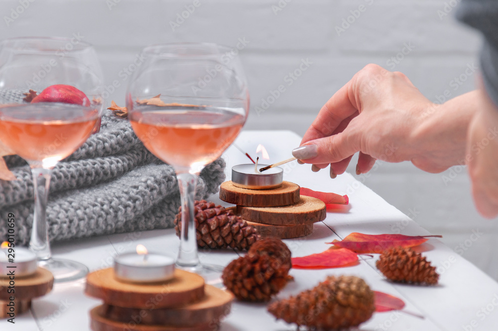 Small burning candles, two glasses with rose wine, cones, dry red leaves, gray scarf knitted on a white wooden table. Girl lights candles on the table. Romantic evening. Hello, Autumn. Cozy autumn bac