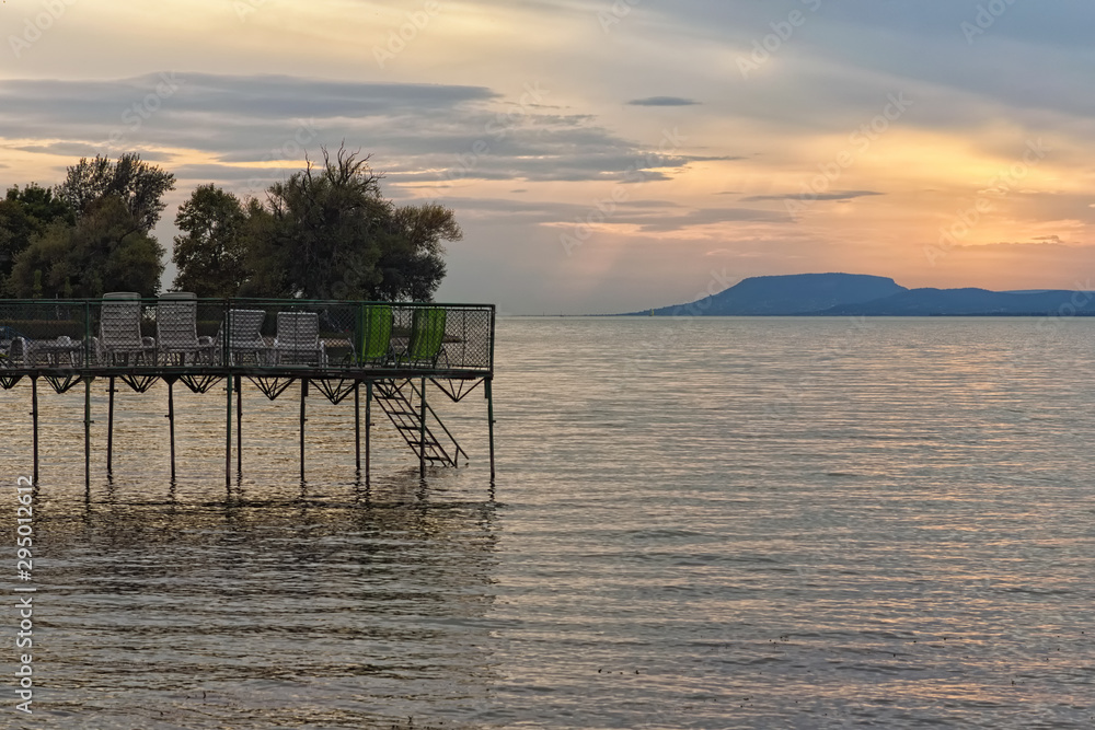 Pier with empty sun loungers at the Lake Balaton during sunset. Sun reflected in the water, mountain at the background. Stunning autumn landscape. Balatonfoldvar, Hungary