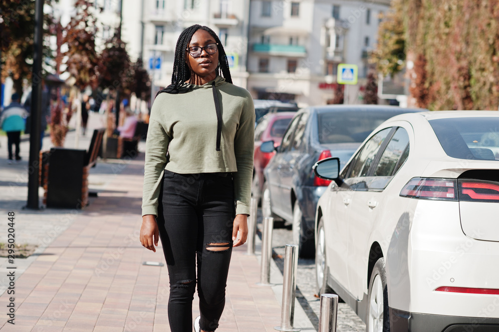 City portrait of positive young dark skinned female wearing green hoody and eyeglasses walking at car parking. City portrait of positive young dark skinned female wearing green hoody and eyeglasses walking at car parking.