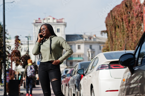 City portrait of positive young dark skinned female wearing green hoody and eyeglasses walking at car parking with mobile phone.