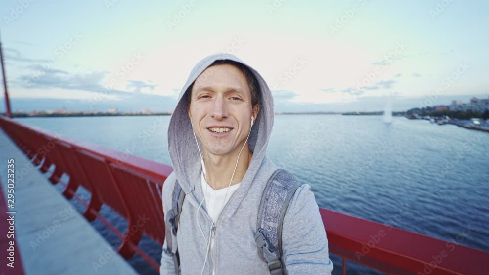 attractive smiling young man Travel Tourist blogger in hoodie making video selfie on the bridge on the big river ang sunset sky background. slow motion fullHD stock footage attractive smiling young man Travel Tourist blogger in hoodie making video selfie on the bridge on the big river ang sunset sky background. slow motion fullHD stock footage