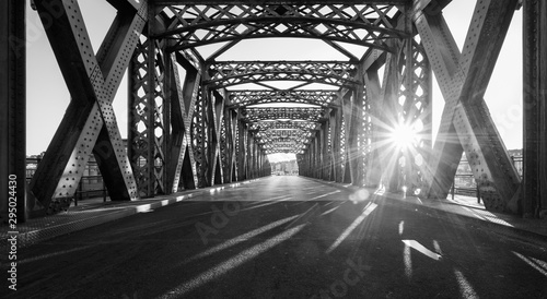 Fototapeta Naklejka Na Ścianę i Meble -  Black and white asphalt road under the steel construction of a bridge in the city on a sunny day. Evening urban scene with the sunbeam in the tunnel. City life, transport and traffic concept.	