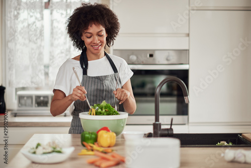Attractive mixed race woman in apron and with curly hair mixing vegetables in bowl while standing in kitchen at home.