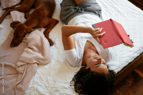 Young woman with dog lying on bed while reading book