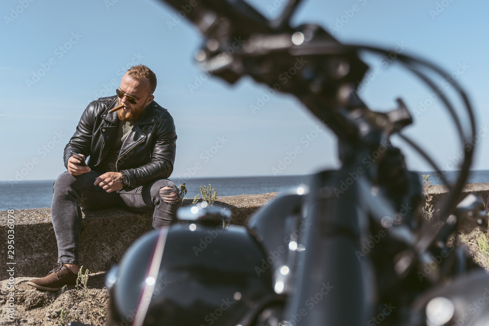 Serious confident man in leather jacket sitting at the roadside with his bike near