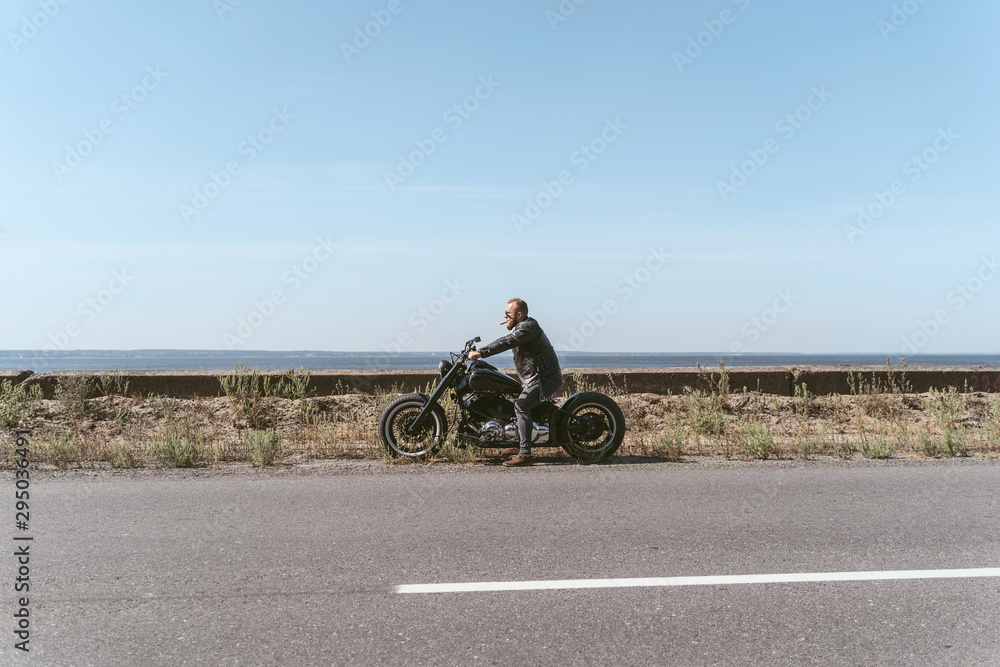 Brutal man in leather jacket and sigar sitting on bike near ocean