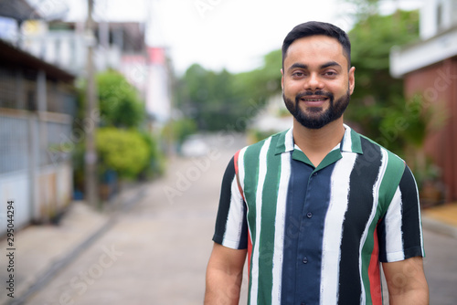 Happy young handsome bearded Indian man smiling outdoors