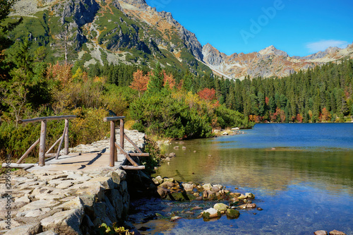 Fototapeta Naklejka Na Ścianę i Meble -  Wooden bridge and stone walkway at the  autumn Poprad lake surrounded by peaks and colorful forests in High Tatras National Park, Slovakia