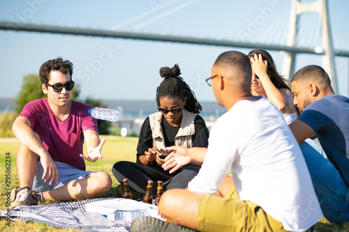 Multiethnic group of friends enjoying dinner outdoors. Young men and woman sitting on grass at plaid with beer and pizza. Dinner outside concept