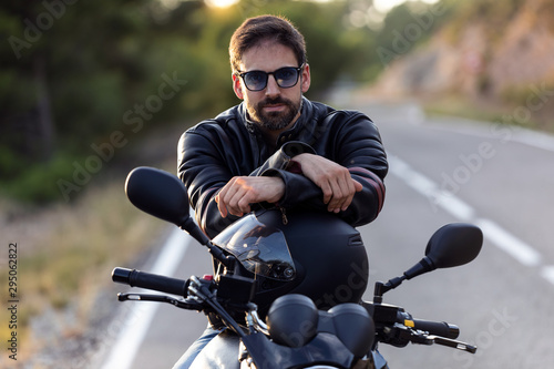 Tableau sur toile Handsome bearded biker looking at camera while sitting on his motorbike and holding helmet
