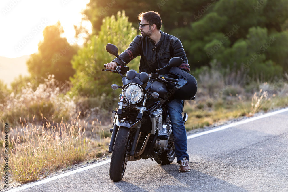 Fototapeta premium Handsome bearded biker looking to sideways while sitting on his motorbike and holding helmet.