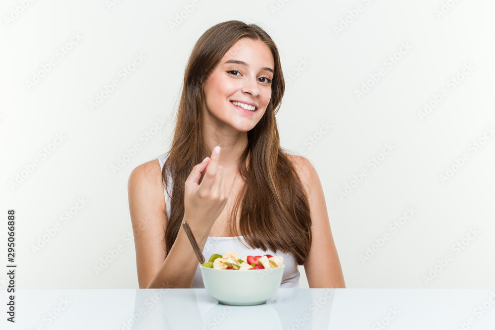 Young caucasian woman eating fruit bowl pointing with finger at you as if inviting come closer. Young caucasian woman eating fruit bowl pointing with finger at you as if inviting come closer.