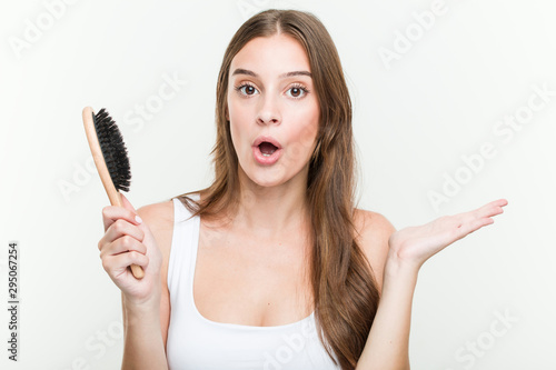 Young caucasian woman holding a hair brush impressed holding copy space on palm.