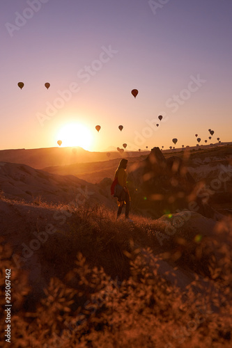 sunrise photo in Cappadocia with air balloons in the sky over sandy hills and a girl standing on the ground
