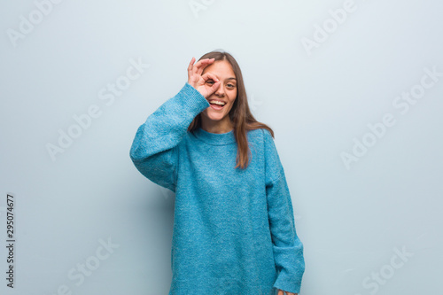 Young pretty woman wearing a blue sweater confident doing ok gesture on eye