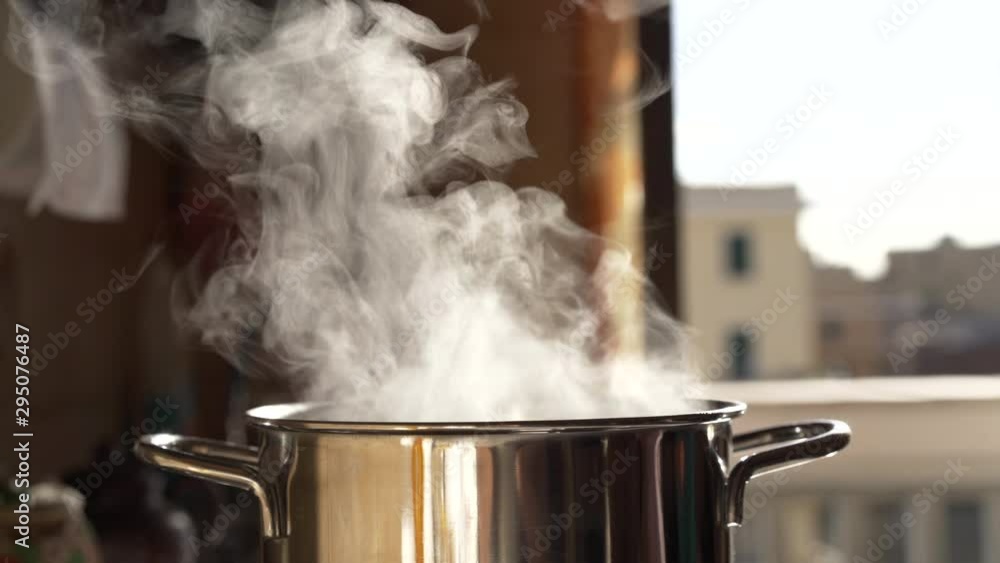 Steaming and boiling pan of water on modern heating stove in kitchen on the background of open balcony. Boiling with steam emitted from stainless cooking pot. Evaporation effects in the air Steaming and boiling pan of water on modern heating stove in kitchen on the background of open balcony. Boiling with steam emitted from stainless cooking pot. Evaporation effects in the air