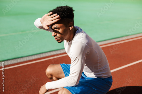 Young thoughtful African American sportsman sit on one's haunches covering face from sun at stadium