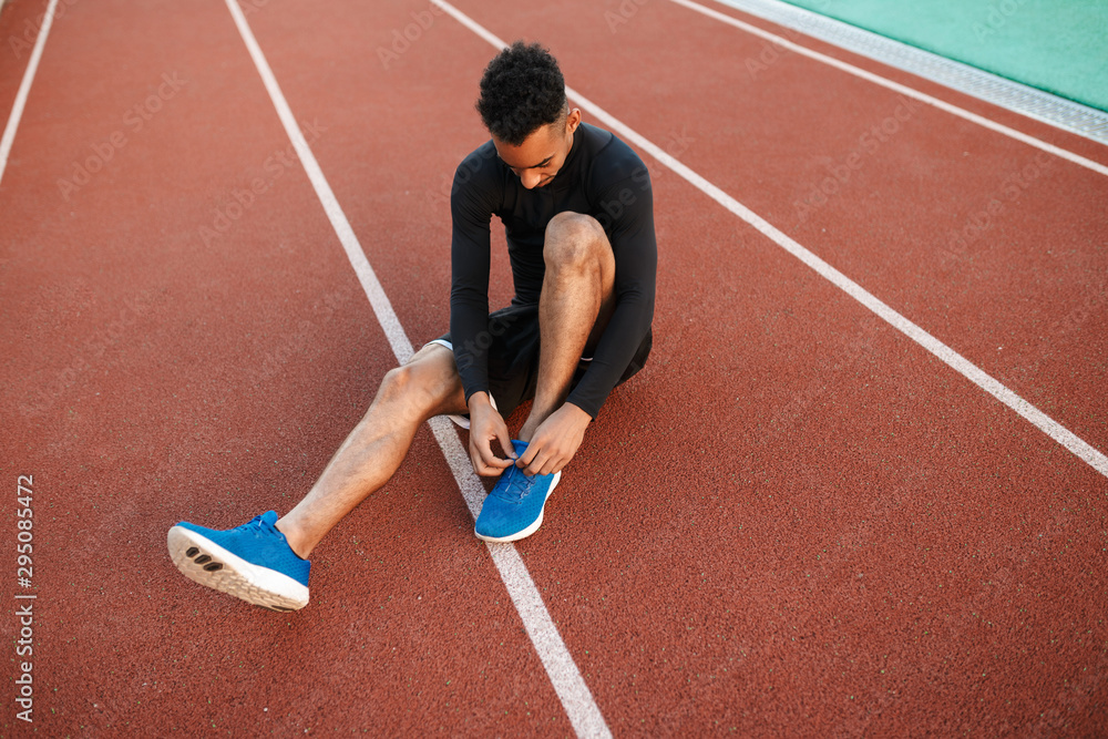 Young African American sporty man tying shoelaces on sneakers on racetrack at city stadium