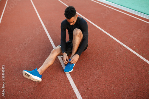 Young African American sporty man tying shoelaces on sneakers on racetrack at city stadium