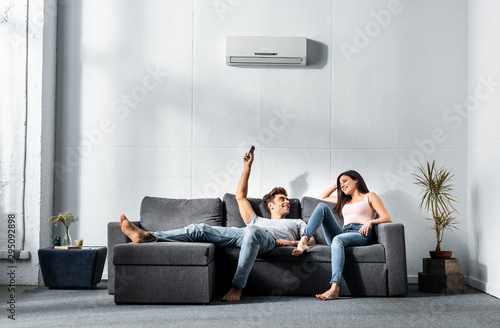 Fotografi handsome boyfriend switching on air conditioner and looking at smiling girlfrien