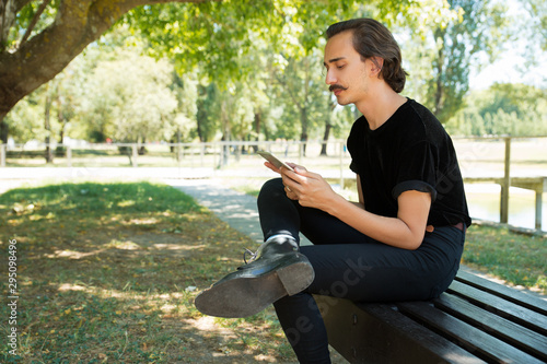 Peaceful serious guy reading on tablet screen outdoors. Young man with curly moustache sitting on park bench, holding and using digital device, looking at screen. Wi-Fi outside concept