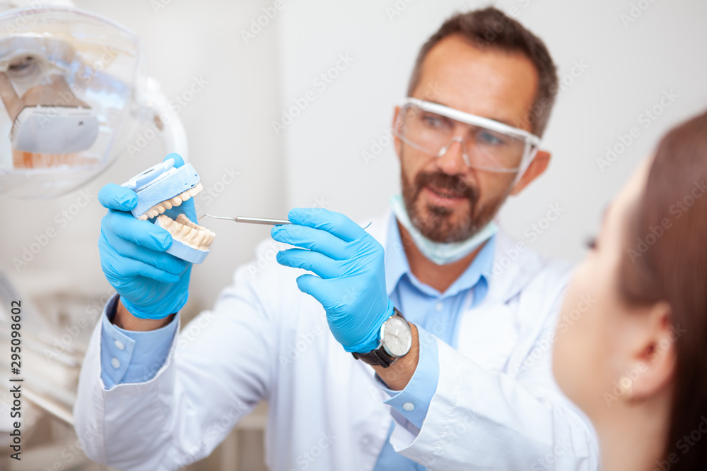 Experienced mature male dentist explaining dental hygiene to his female patient, holding teeth mold Experienced mature male dentist explaining dental hygiene to his female patient, holding teeth mold