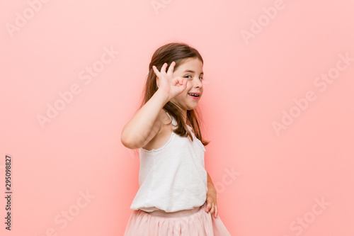 Little girl wearing a princess look winks an eye and holds an okay gesture with hand.