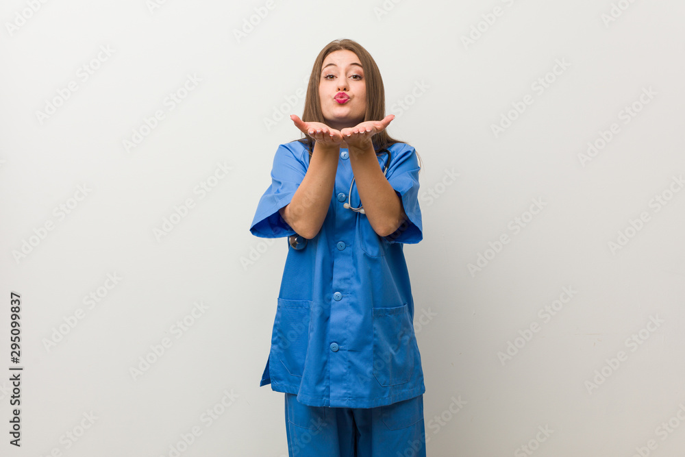 Young nurse woman against a white wall folding lips and holding palms to send air kiss. Young nurse woman against a white wall folding lips and holding palms to send air kiss.