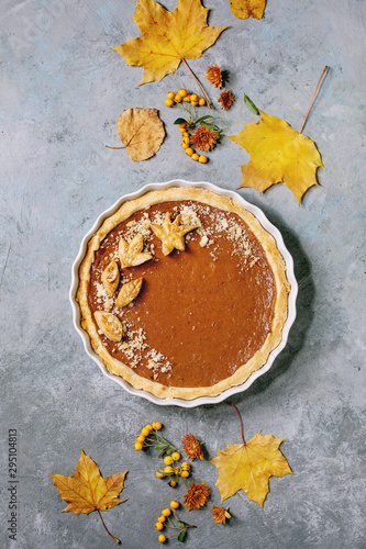 Traditional homemade autumn pumpkin pie for Thanksgiving or Halloween dinner served in ceramic dish with yellow autumn leaves over grey texture background. Flat lay, space
