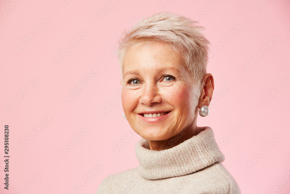 Portrait of mature woman with short blond hair and in beautiful earrings smiling at camera over pink background Portrait of mature woman with short blond hair and in beautiful earrings smiling at camera over pink background