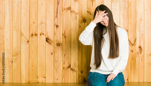 Young chinese woman sitting on a wooden place blink at the camera through fingers, embarrassed covering face. © Asier