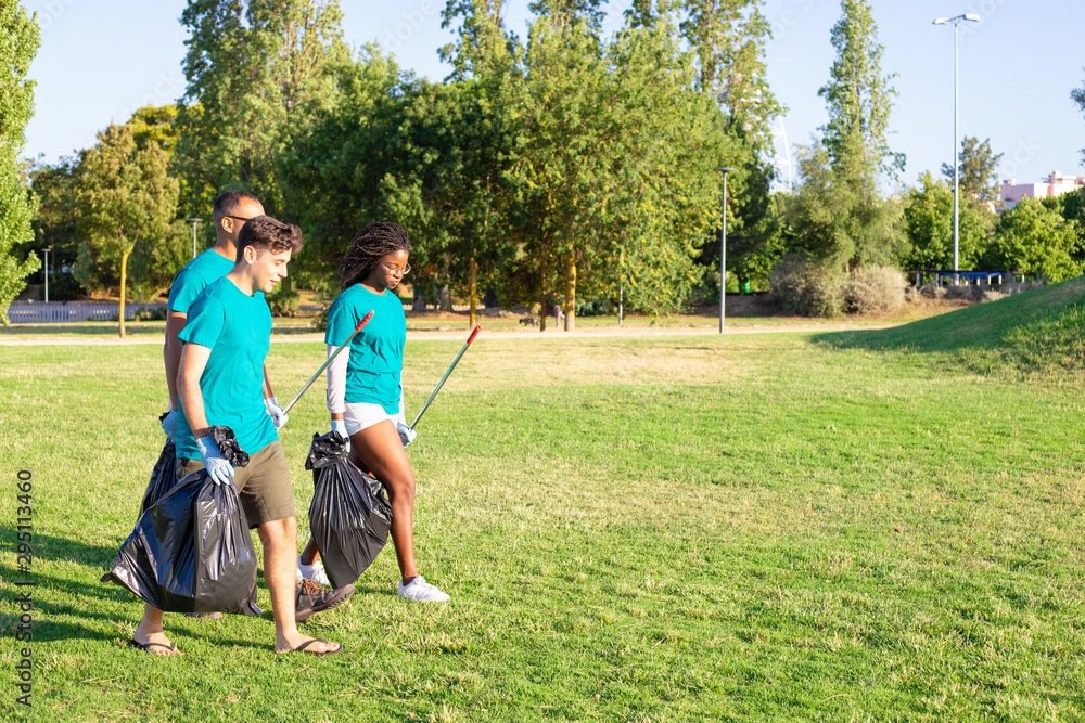 Team of eco activists leaving park after cleaning lawns. Young men and woman walking on grass, carrying rakes, plastic bags and talking. Garbage collection concept