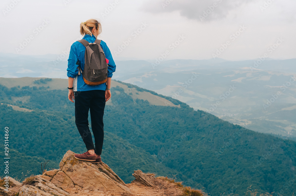 Young girl tourist with  backpack behind her stands on top of the mountain and enjoys a view of the Carpathian mountains at sunrise.