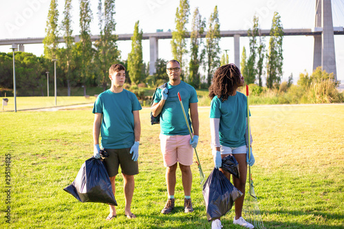 Eco activist team cleaning city park. Young men and woman standing on grass, holding rakes, plastic bags, looking around. Garbage removal concept photo