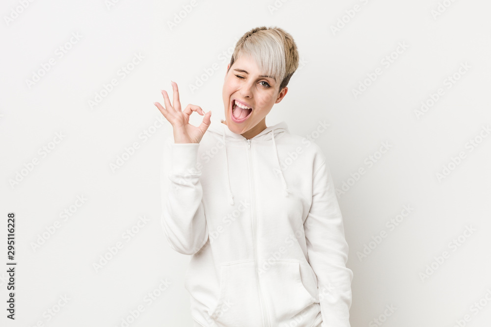Young curvy woman wearing a white hoodie winks an eye and holds an okay gesture with hand. Young curvy woman wearing a white hoodie winks an eye and holds an okay gesture with hand.