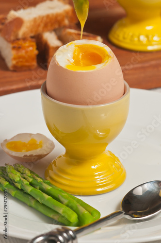 Top, front view, close up of a brown egg in a yellow porcelain egg holder with top cut off showing soft yolk with top on side and asparagus on a white round plate