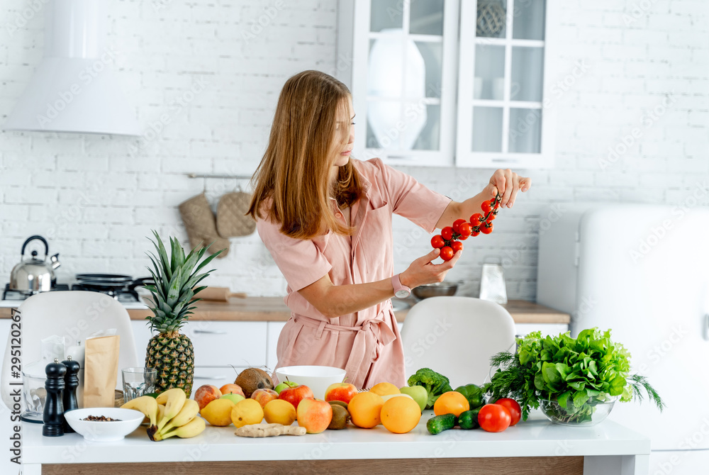 Woman in kitchen ready to prepare meal with vegetables and fruits. Woman is looking at tomatoes in her hands. Kitchen background. Healthy food. Vegans. Vegeterian.