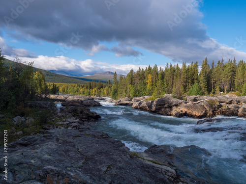 Beautiful northern landscape with wild glacier river Kamajokk, boulders and spruce tree forest in Kvikkjokk in Swedish Lapland. Summer sunny day, golden hour, dramatic clouds