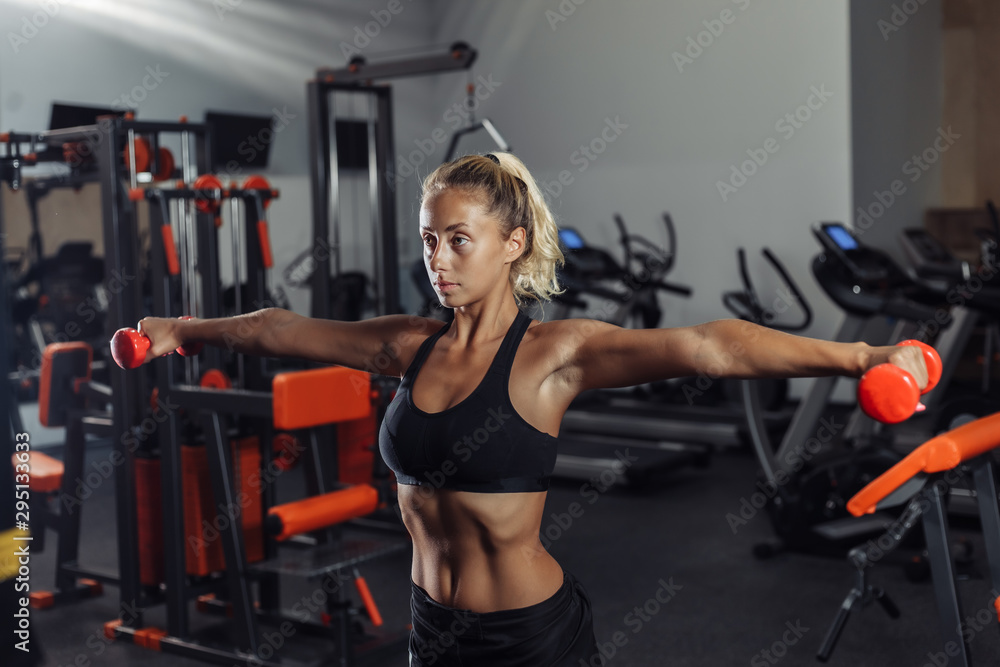 Fototapeta premium Young fit woman in sportswear is training with dumbbells in the gym. Healthy lifestyle concept. Body training with free weights