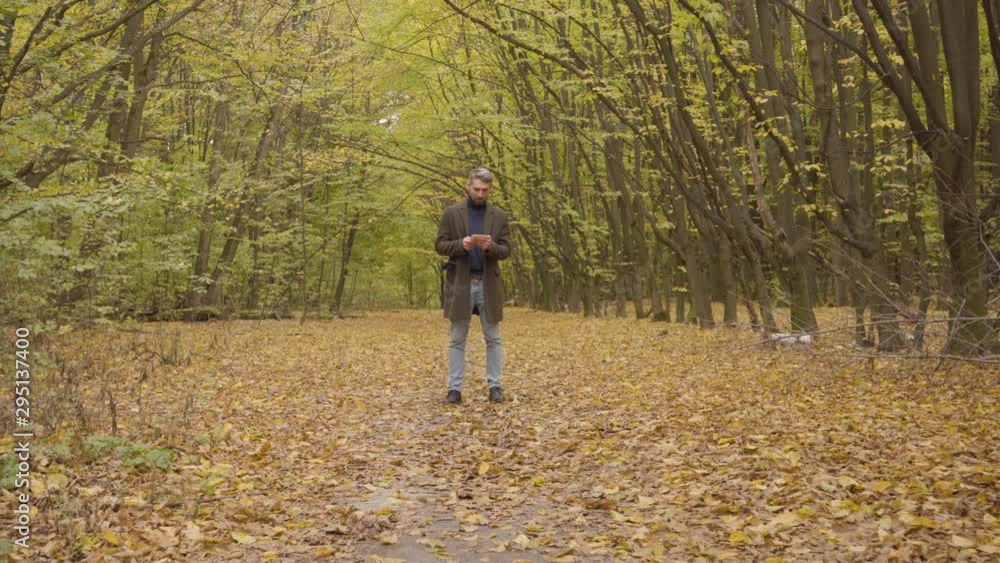 Camera approaching to a confident handsome man standing in the autumn forest and using his tablet. Serious Caucasian businessman working on weekends outdoors. Camera approaching to a confident handsome man standing in the autumn forest and using his tablet. Serious Caucasian businessman working on weekends outdoors.